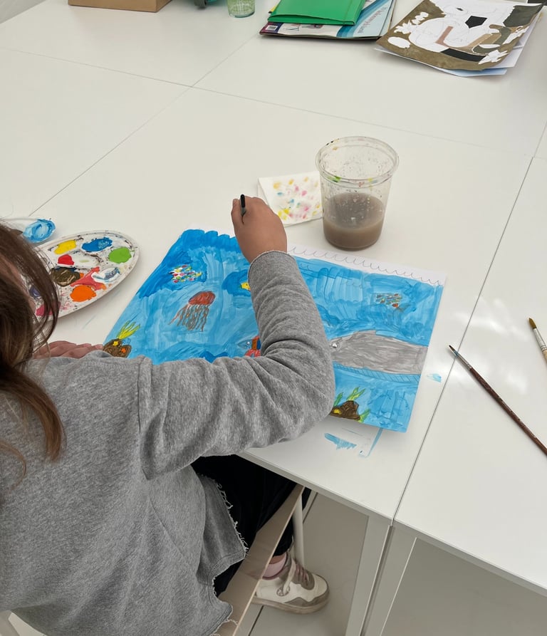 A kid at a desk painting a blue ocean scene with jellyfish and sea creatures.
