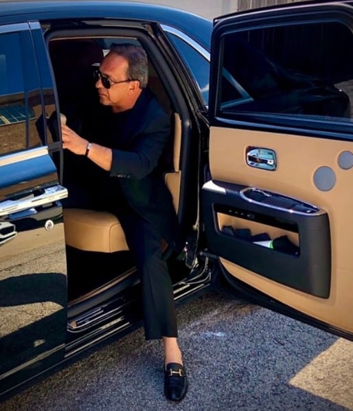 A stylish man, Bedig Minassian, in a dark suit and sunglasses, steps out of a classic car