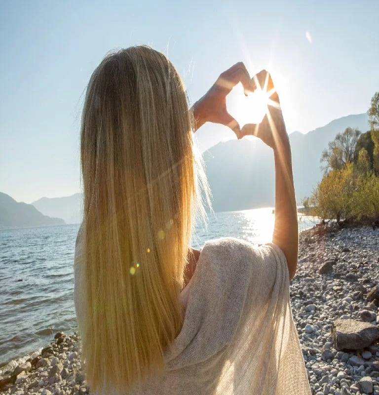 Orlane making a heart shape with her hands against a bright sunset over a mountain lake.