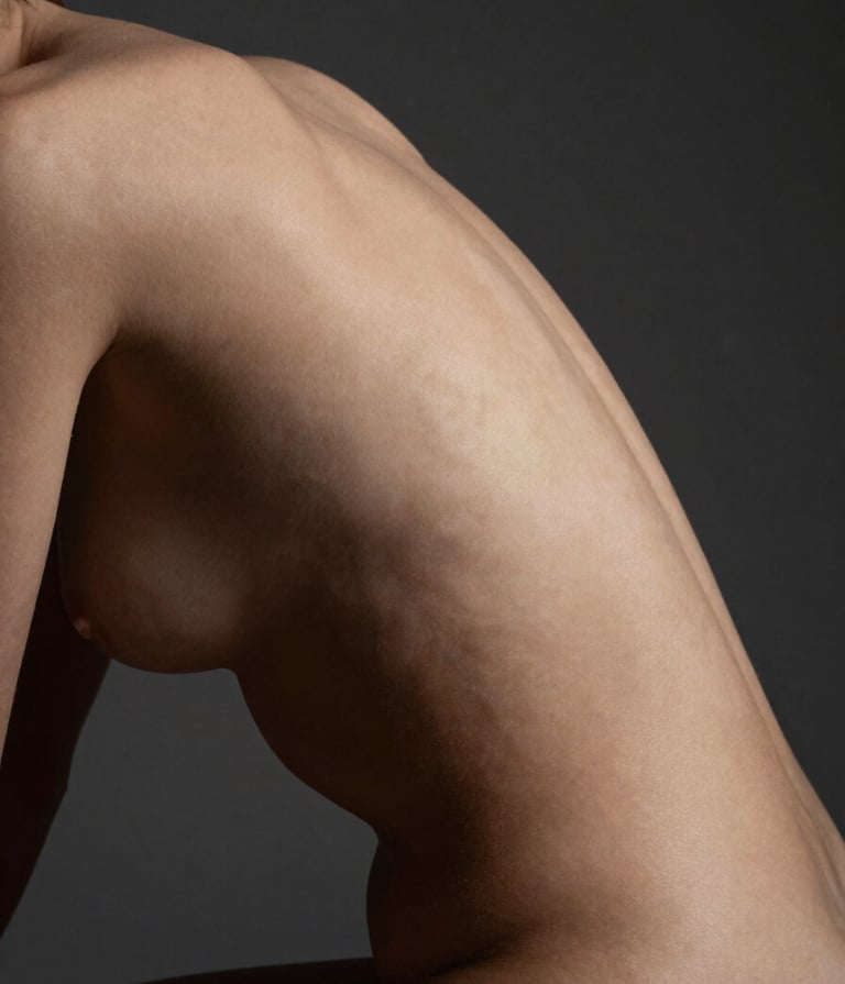 A sophisticated fine-art nude photograph focusing on the soft curves of a back and spine. Minimalist composition with a dark gray background. Lighting is soft and directional, creating a gentle gradient. Professional photography, North American / US studio setting.