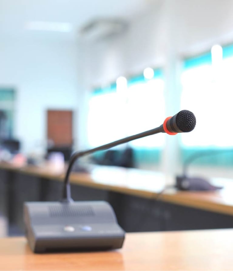 A close-up of a gooseneck conference microphone with a red ring around the mic head, placed on a wooden table in a brightly l