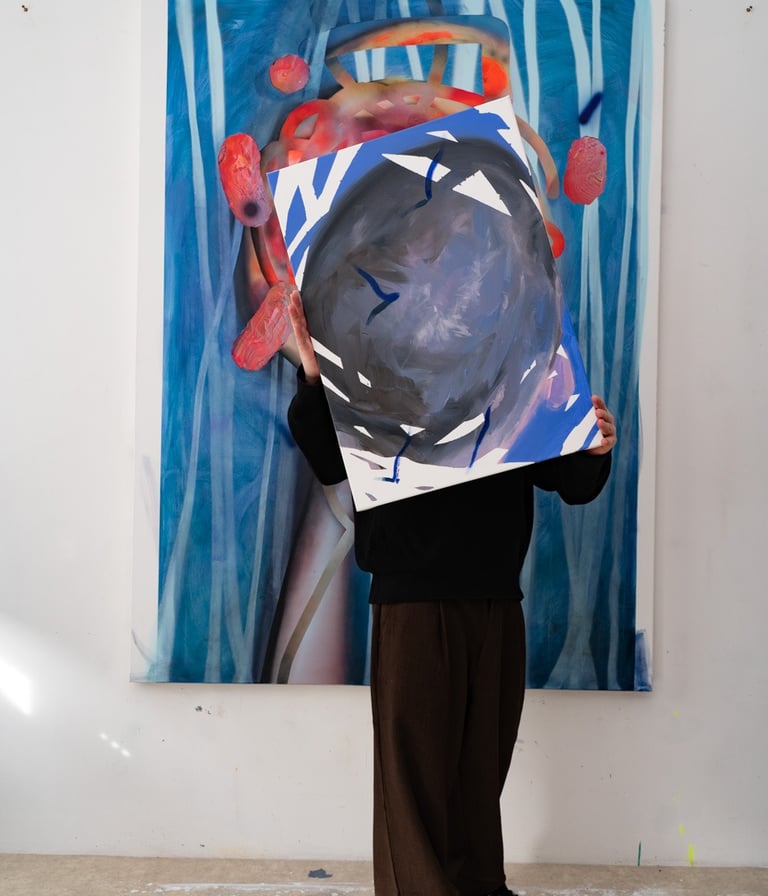 Artist in a studio holding a contemporary abstract painting over their face in front of a larger blue canvas.