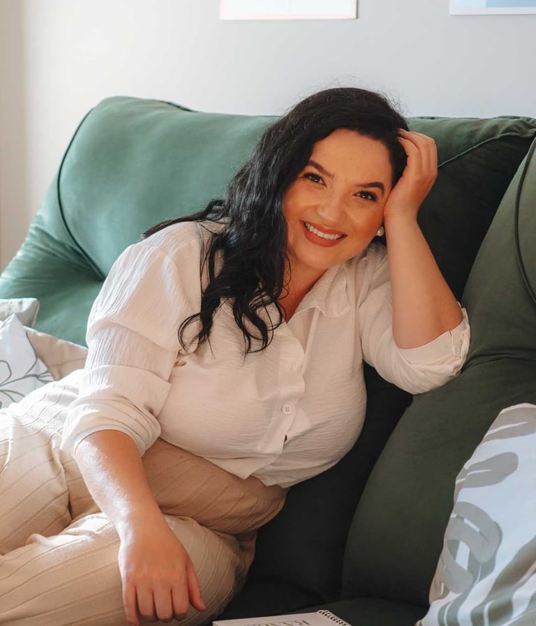 Raabe sentada em sofá verde sorrindo com caderno ao lado.