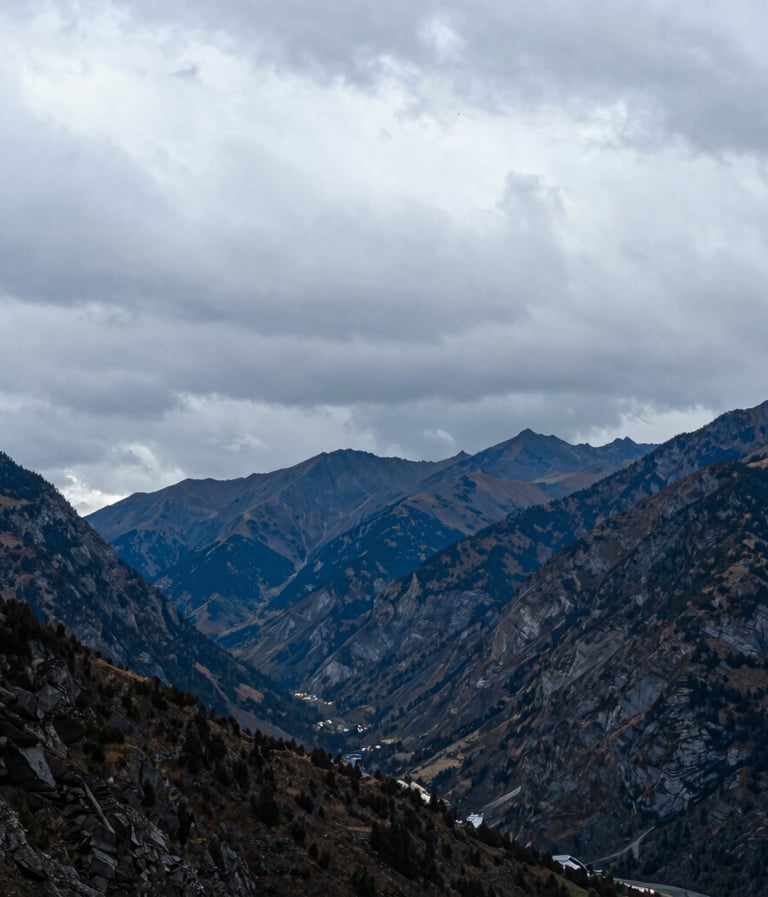 A vertical landscape photograph of an Alpine valley just before a storm. The mountains are deep charcoal blue against a turbulent, light grey sky. The composition is spacious and highlights the grandeur of the Western landscape.