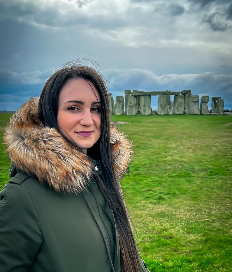 A woman in a green parka smiles in front of the prehistoric Stonehenge monument under a cloudy sky.