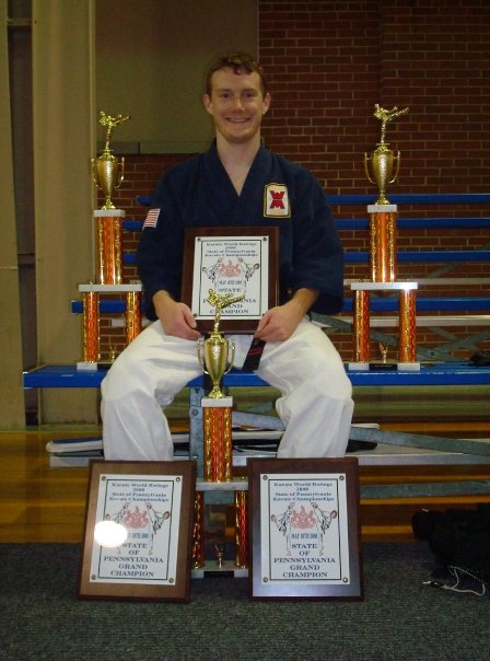 A karate grand champion in a blue gi sits with his martial arts trophies and awards.