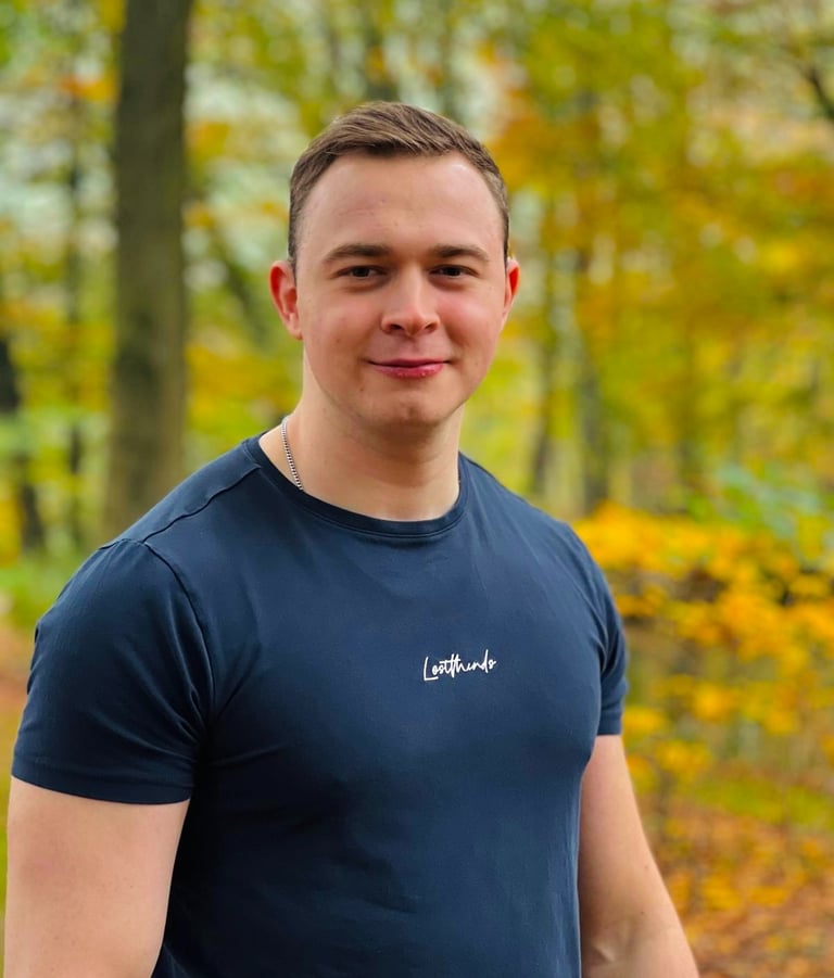 A smiling man in a navy blue t-shirt stands outdoors against a background of yellow autumn foliage.