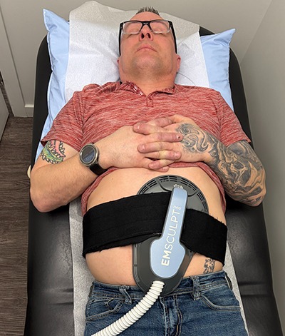 A patient in a doctors office laying on a bed using the BLT Emsculpt on his abdomen 