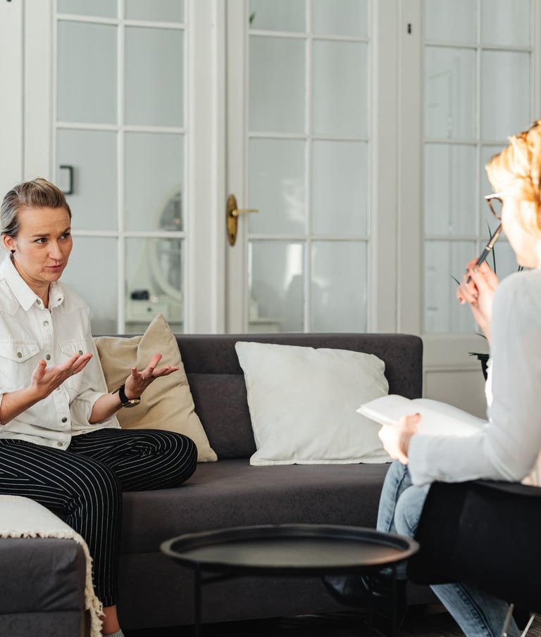 Two women in a therapy session, one gesturing while speaking, the other taking notes