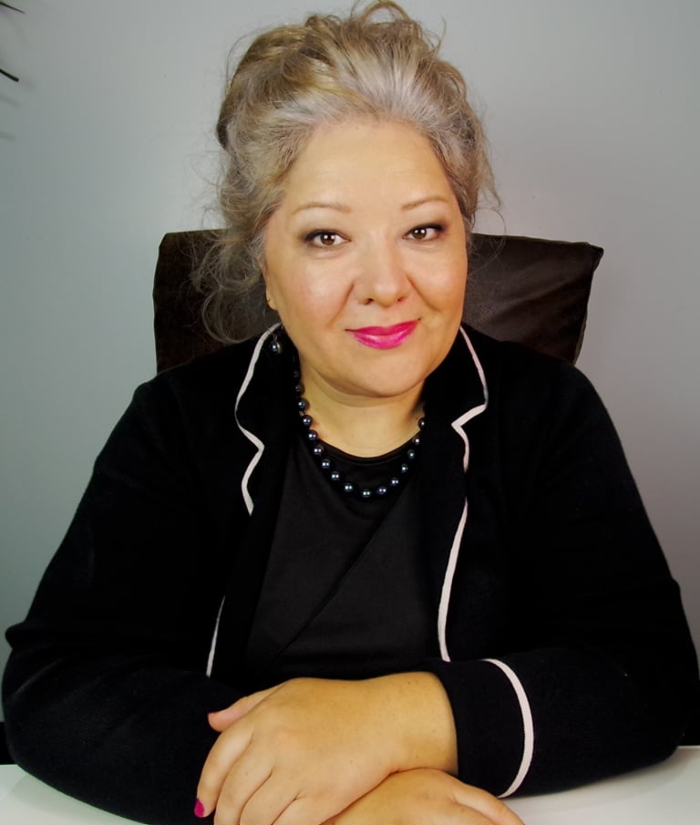 Professional woman with grey hair and black blazer sitting at a white desk in an office.