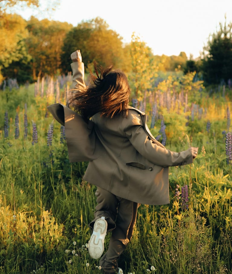 a woman in a coat and sneakers is running through the grass her left arm raised boldly