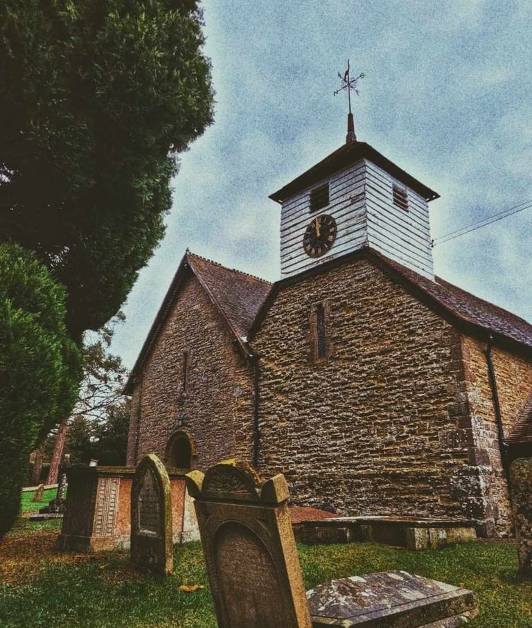 Historic stone country church with a clock tower and graveyard under a cloudy sky.