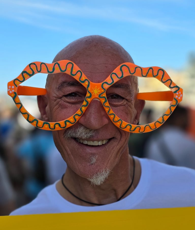 Older man smiles, wearing big colorful lips-shaped glasses outdoors, blue sky above and a blurred crowd behind him.