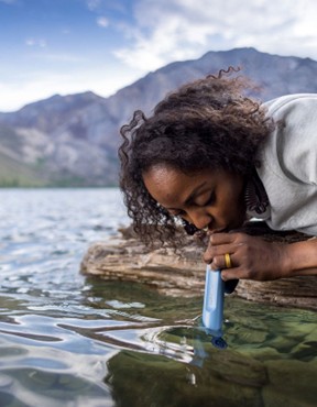 A hiker uses a portable water filter straw to drink from a mountain lake while camping.