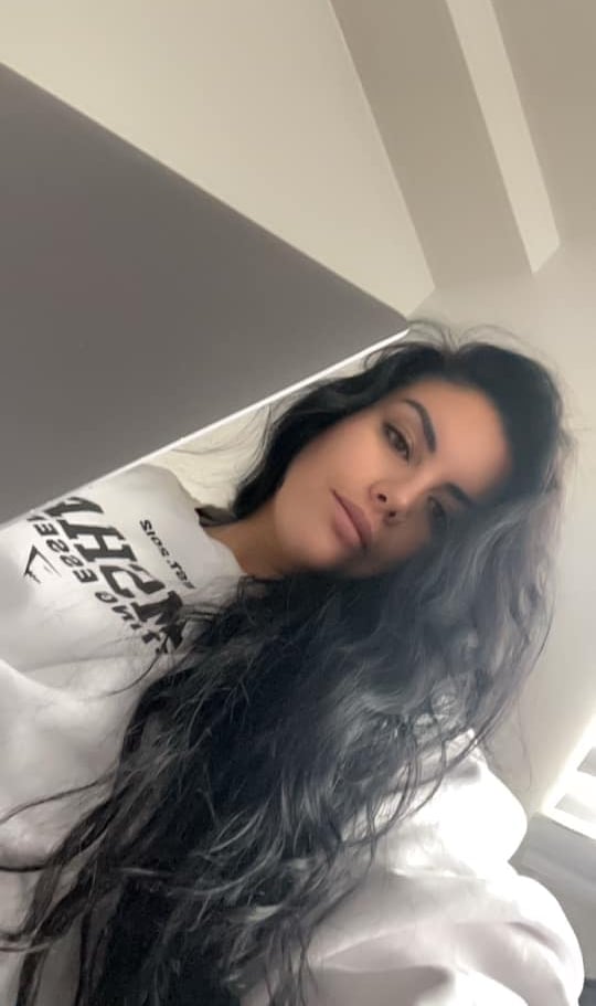 Young woman with long dark wavy hair wearing a white sweatshirt looking into a laptop camera.