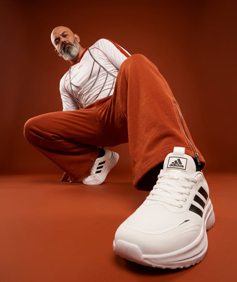 A man in orange streetwear wearing white Adidas running shoes with black stripes against a brown background.