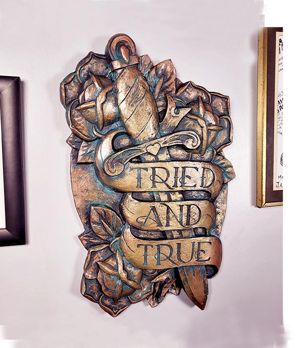 A plaque in the style of traditional tattoo art. Dagger with roses and a banner saying Tried & True