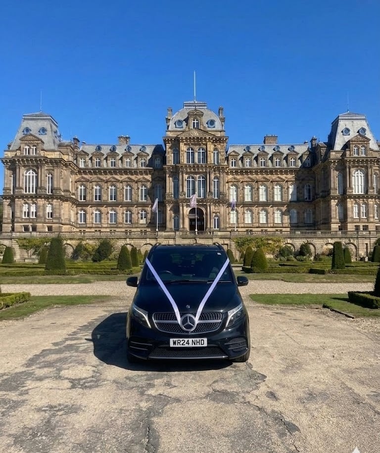 Black Mercedes wedding car with white ribbons parked in front of a grand historic manor house estate.