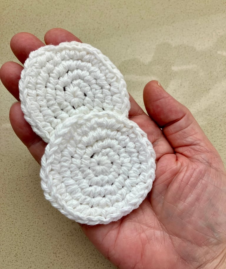 Hand holding 2 small, crocheted, white, round, face scrubbies