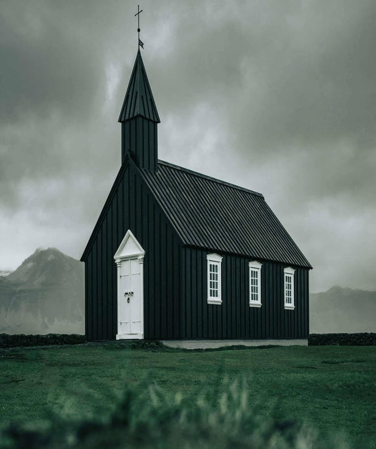 Small black church with white trim and cross-topped steeple set against dramatic cloudy sky.