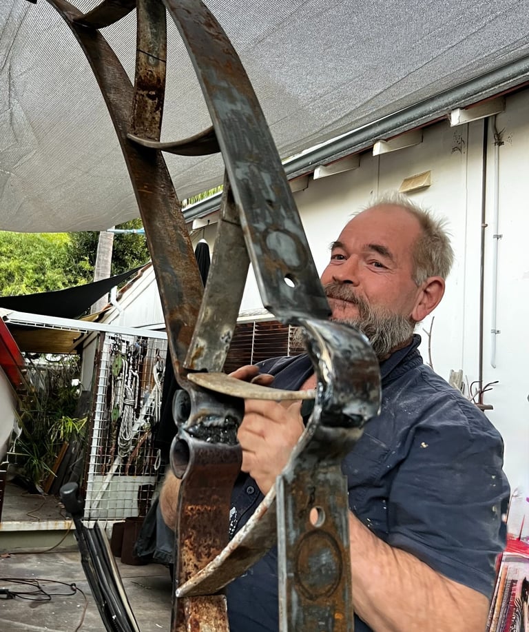 A bearded metal artist working on a large, curved steel sculpture in an outdoor workshop.