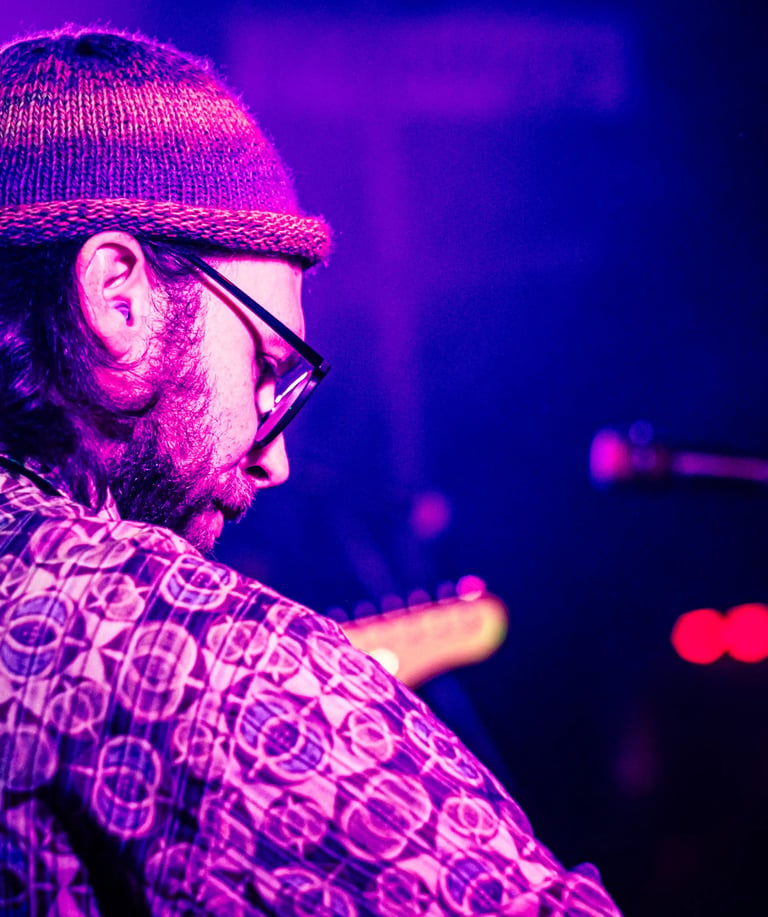 A bearded musician with a beanie plays electric guitar during a live concert with purple stage lighting.