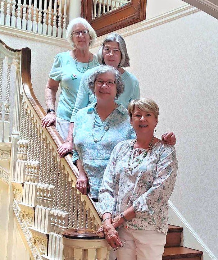 Four smiling senior women posing together on a historic wooden staircase in a bright home.