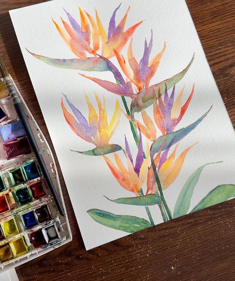 Vibrant watercolor painting of a Strelitzia flower with orange and blue petals resting on an artist's workspace.