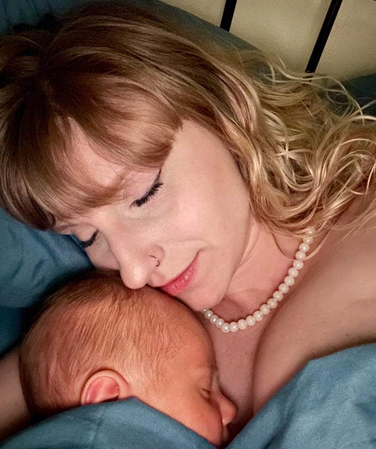 A mother with blonde hair and a pearl necklace peacefully cuddles her sleeping newborn baby, both wrapped in a blue blanket