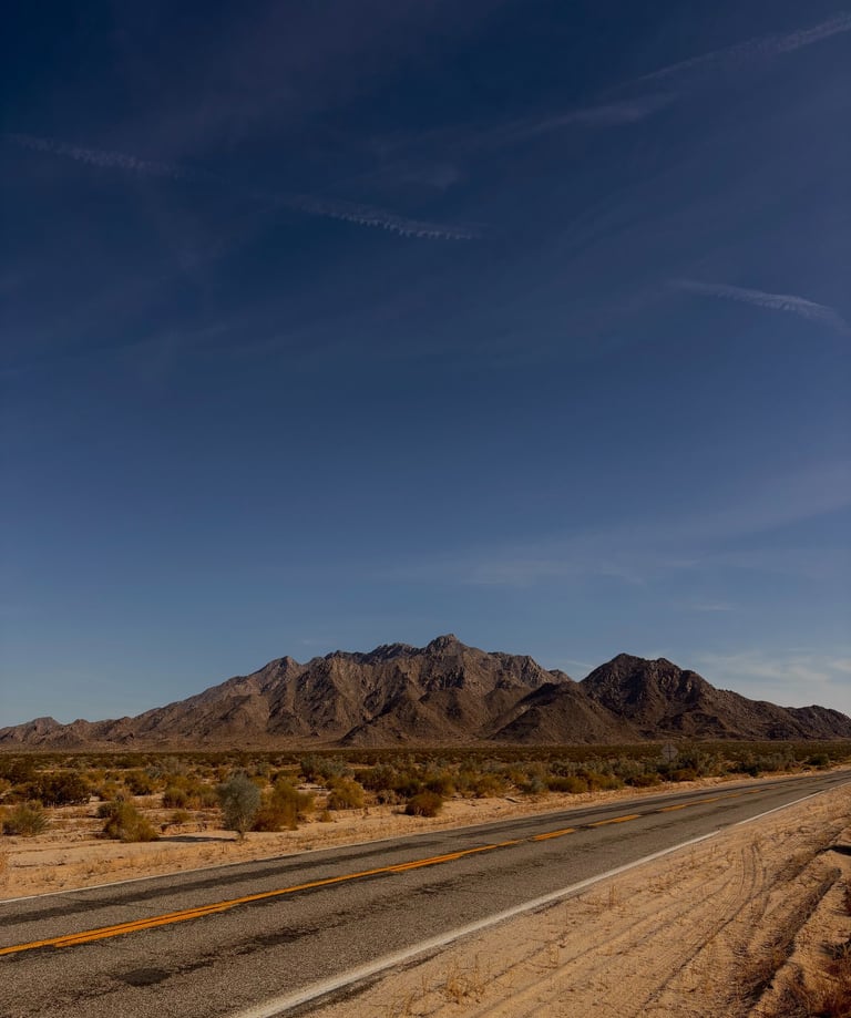 A California desert highway leading toward rugged mountains under a clear blue sky.