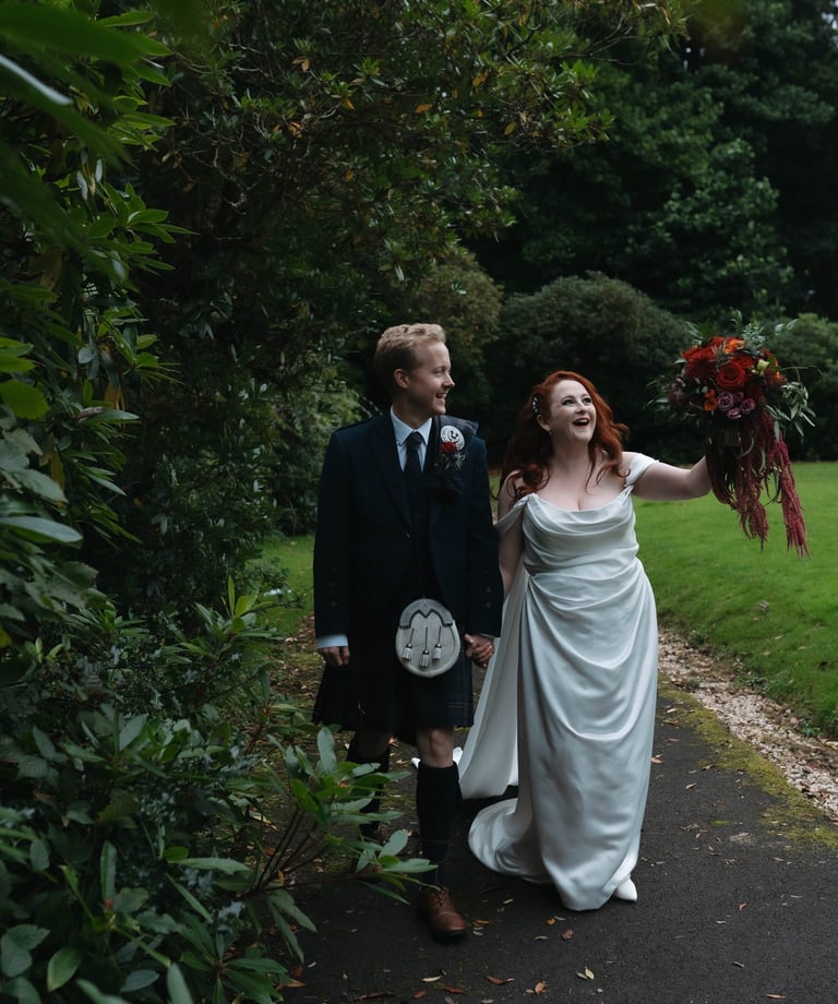 A bride in a white wedding gown and groom in a traditional Scottish kilt walk through a lush garden path.