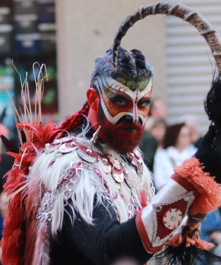 Man in elaborate red and white tribal costume with face paint and horns at a festival.