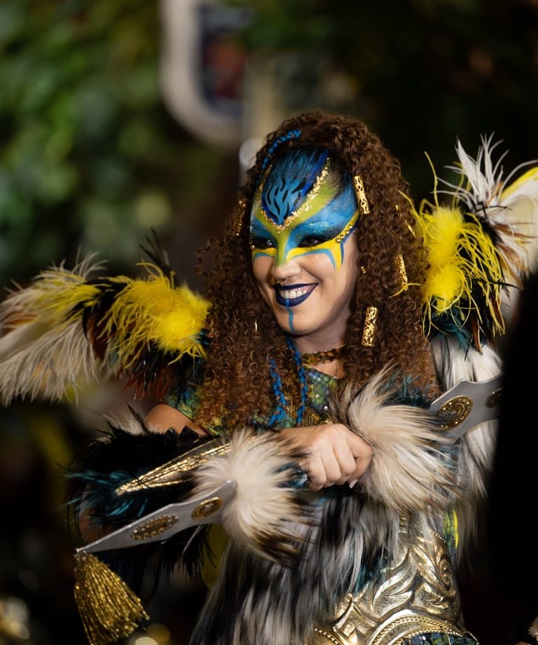 Smiling woman in fantasy warrior costume with blue face paint, feathers, and golden armor.