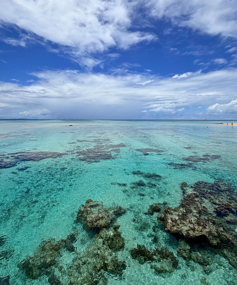 Clear turquoise tropical ocean water with coral reefs under a blue sky with white clouds.