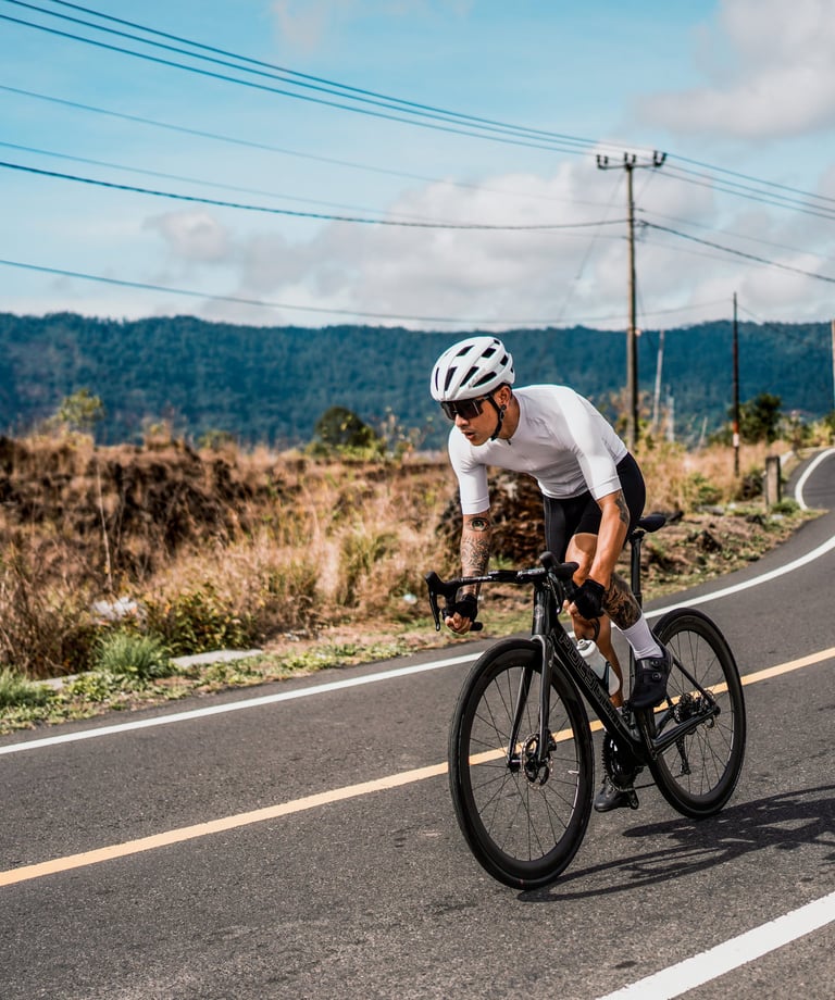 A cyclist in white gear riding a black road bike on a winding mountain highway.