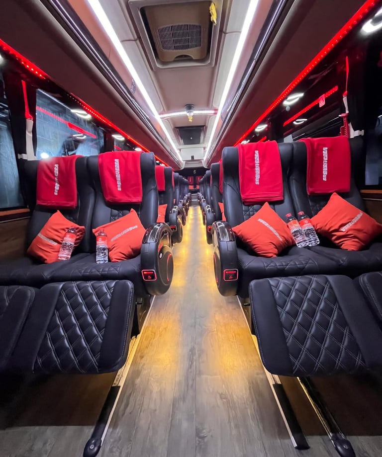 Luxury tour bus interior featuring black leather recliner seats, orange pillows, and red ambient lighting.