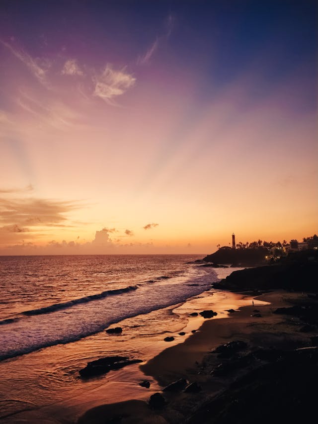 A scenic sunset over a coastal beach featuring a distant lighthouse on a cliff and purple sky.