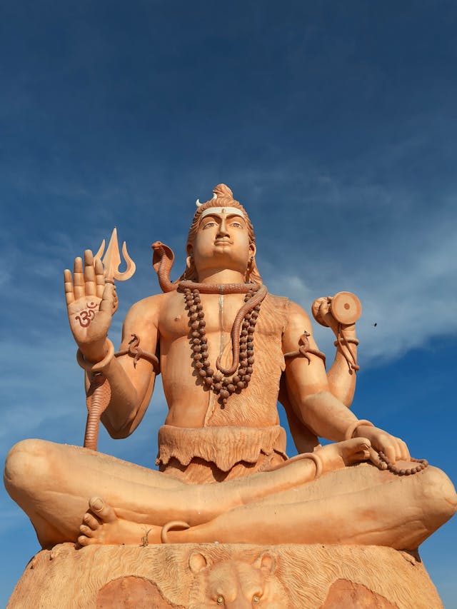 A tall sandstone statue of Hindu deity Lord Shiva in a meditative pose against a clear blue sky.