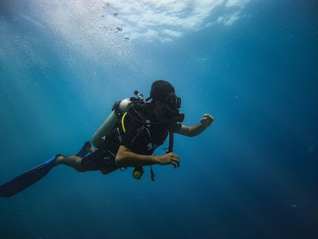 A scuba diver swimming through deep blue ocean water with sun rays and air bubbles.