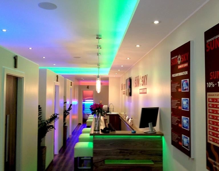 Zabudowa w salonie opalania z kolorowym podświetleniem LED, drewnianym blatem recepcji i nowoczesnym sufitem z punktowym oświ