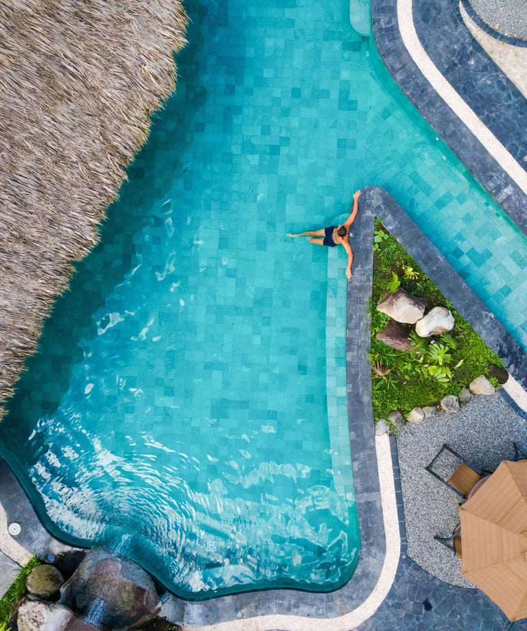 Aerial view of a woman relaxing in a luxury tropical resort swimming pool next to a thatched roof hut.