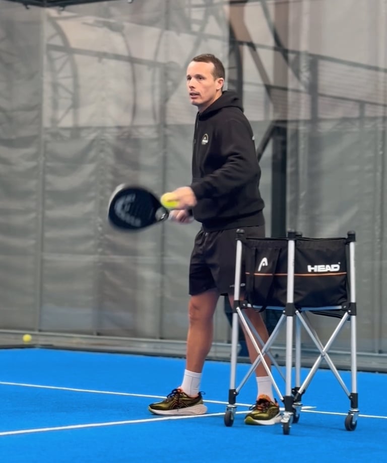 A man practicing padel on a blue indoor court with a racket and a Head ball cart.