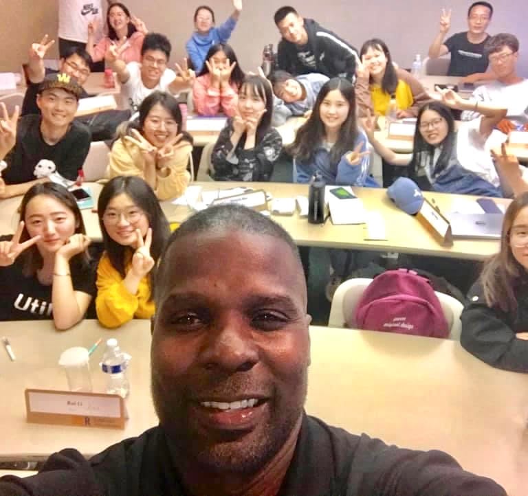 Dr. Marlon Ware standing in front of his class with students holding up the peace sign