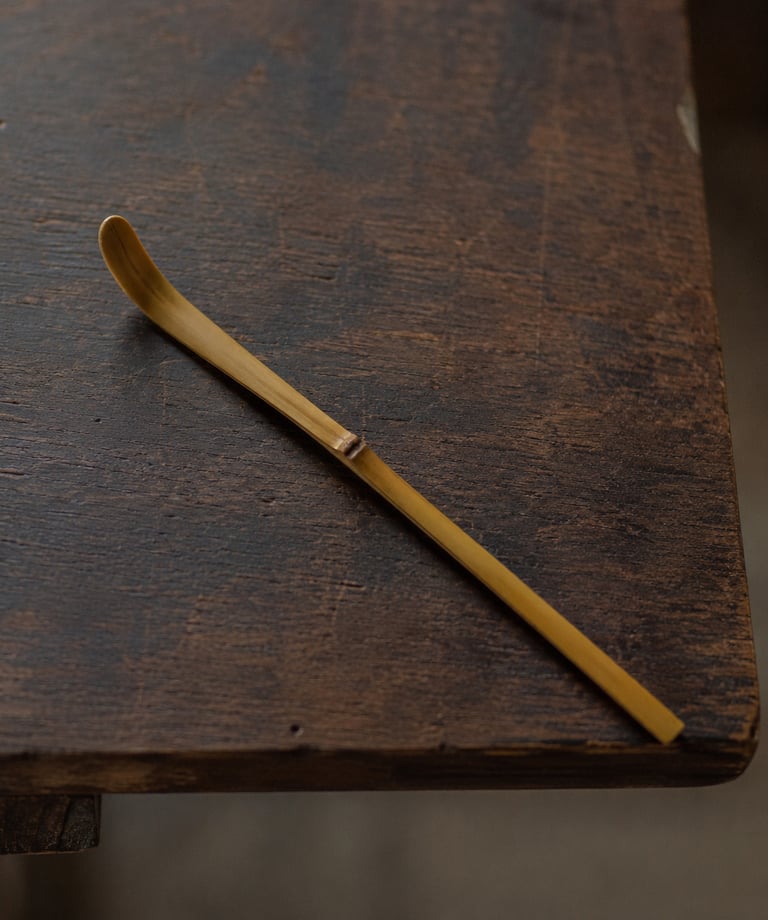 Handcrafted bamboo chashaku matcha tea scoop resting on a rustic dark wood table.