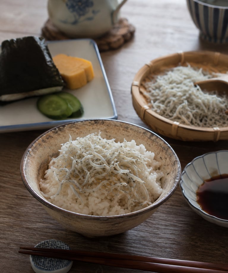 A traditional Japanese breakfast with a bowl of shirasu whitebait over steamed rice, onigiri, and tamagoyaki.