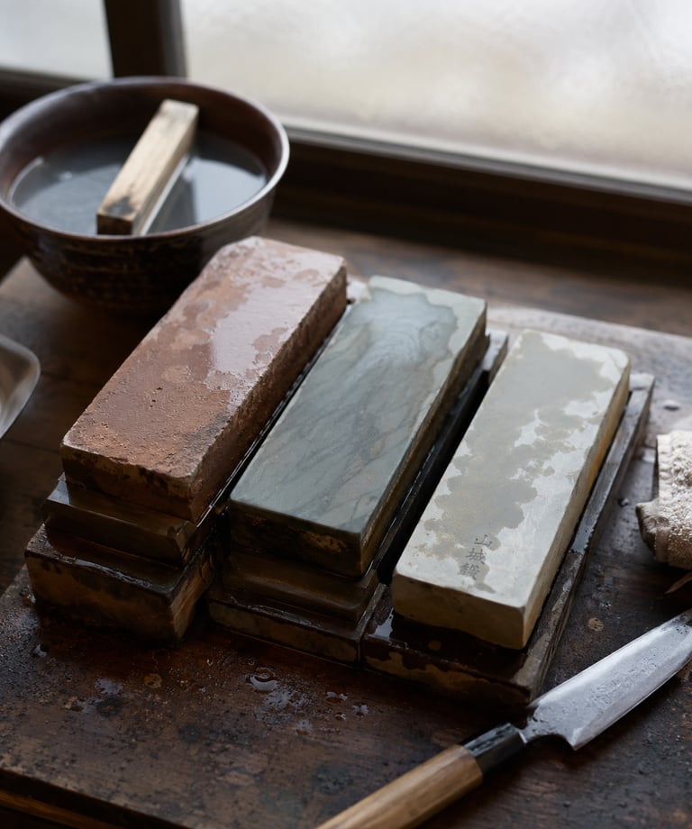A collection of wet Japanese natural whetstones and a chef knife on a wooden workbench for professional sharpening.