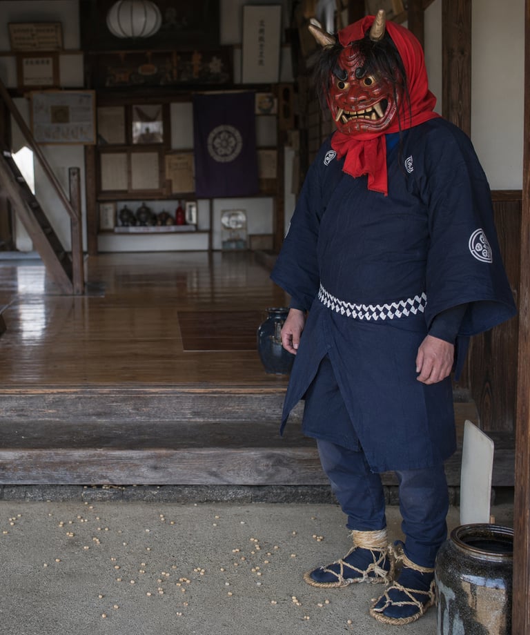 A man wearing a traditional red Japanese Oni demon mask and blue kimono at a temple entrance.