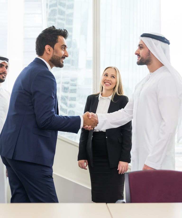 Businessman in a suit shakes hands with another in Emirati attire, while a businesswoman smiles