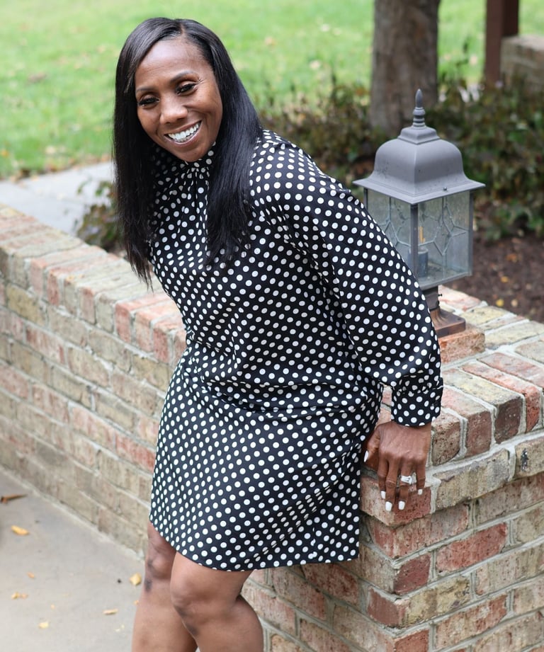Smiling woman in a black and white polka dot dress posing against a brick wall outdoors.