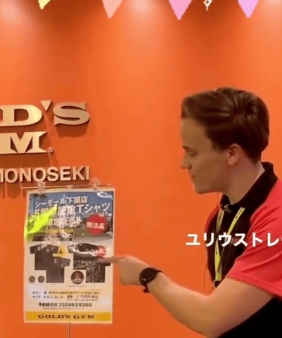 Julius-Sensei steht vor einer orangefarbenen Wand mit dem Gold’s-Gym-Logo und zeigt auf ein Plakat